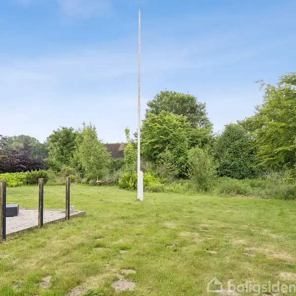 Flagstang står stille på grøn græsplæne omgivet af træer og buske. I venstre side ses en terrasse med flisebelægning og hegn.