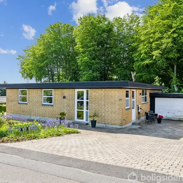 En enkel, gulstensbungalow med fladt tag står stille i en have med farverige blomster, omgivet af tætte grønne træer og en flisebelagt indkørsel.