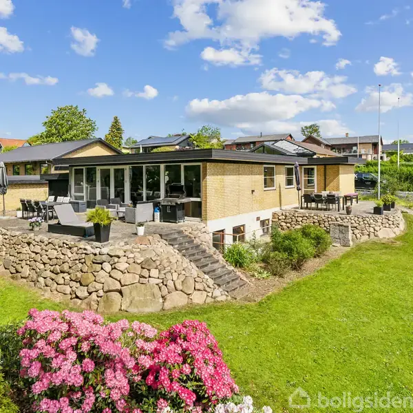 Et gult murstenshus med store vinduer og terrasse står i en velplejet have med grønne græsplæner og blomstrende buske. Omkringliggende ejendomme ses under en skyfri, blå himmel.