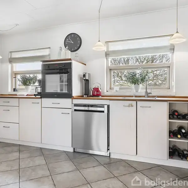 Et moderne køkken med hvide skabe og en indbygget ovn. En opvaskemaskine er ved siden af, mens vinflasker er opbevaret til højre. To vinduer slipper lys ind.