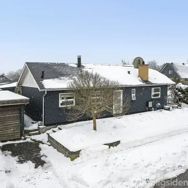 Et sort hus med en sneklædt tag og indgang står ved siden af en carport. Træer og buske omgiver huset i et snedækket nabolag.