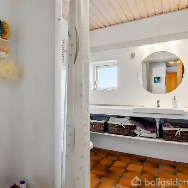 Bruser med hvidt forhæng i et badeværelse. Rundt spejl hænger over dobbelthåndvask med kurve under. Loft af træ og gulv af brune fliser. En dør og væghængte håndklæder.