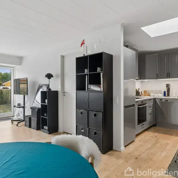 Stue-køkken-alrum med åbent design. Sort reol adskiller områderne, mens vinduer lukker dagslys ind. Rundt spisebord med stole i forgrunden i moderne boligindretning.