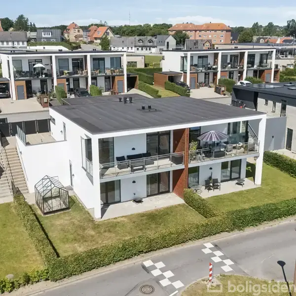 Hvid, moderne to-etagers rækkehus med altan og terrasse; omkranset af græsplæne. Andre lignende boliger i baggrunden og blå himmel. Vej og fortov i forgrunden.