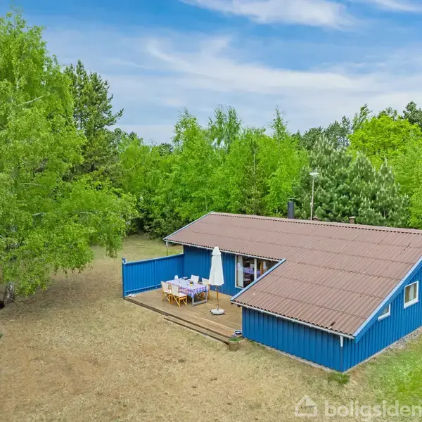 Et blåt træsommerhus står stille blandt grønne træer med en terrasse, hvor der er havemøbler. Solskinnet oplyser området, der virker fredeligt og naturligt.