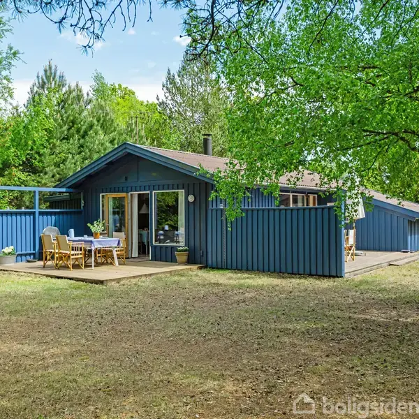 Et blåt træhus med terrasse står stille omgivet af grønne træer. Et bord og stole er placeret på terrassen, som fanger sollyset i den åbne natur.