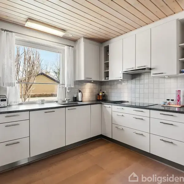 Et hvidt, minimalistisk køkken med sorte bordplader og træloft, hvor apparater som kaffemaskine og mikrobølgeovn står på bordet. Et stort vindue giver udsigt til en have.