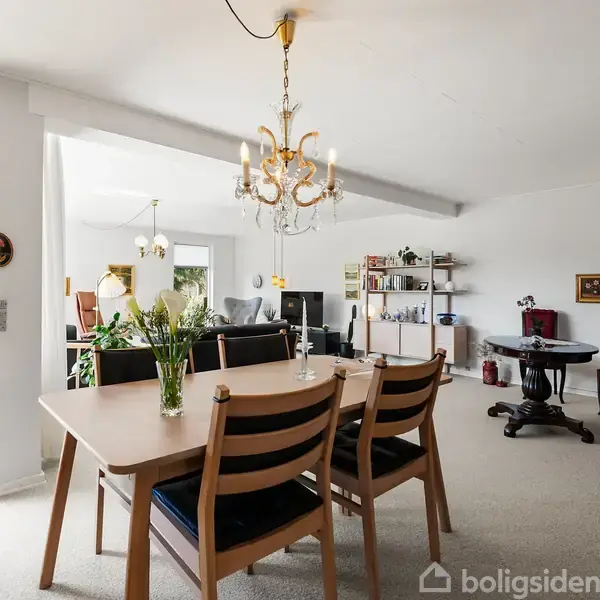 En spisestue med et træbord og seks stole er opstillet med blomster i en vase. Rummet er lyst med store vinduer, en lysekrone og tilstødende opholdsstue med møbler.