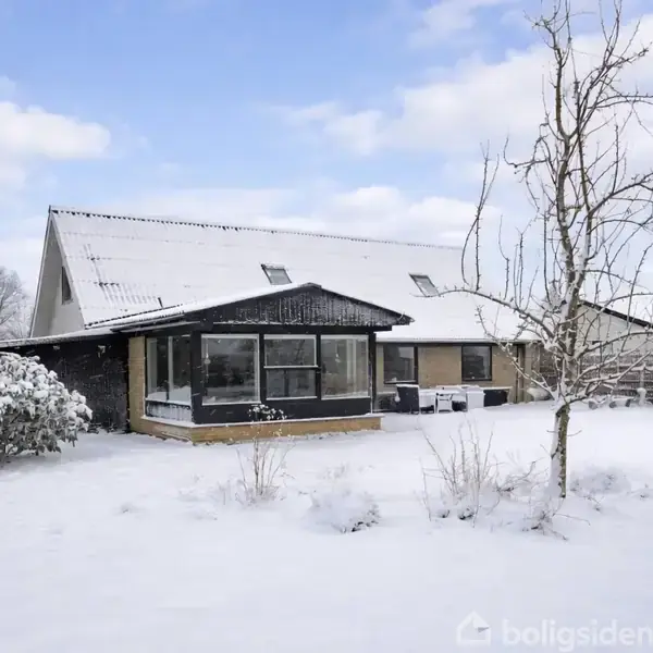 Et hus med store vinduer står i en snedækket have. Træer uden blade omkranser haven, og himlen er blå med nogle skyer i baggrunden.