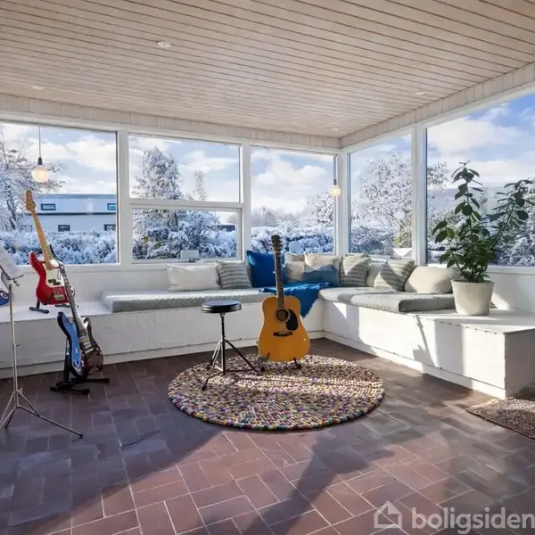 En akustisk guitar står på et rundt tæppe i et solrigt rum med store vinduer. Væggene har flere guitarer ophængt. Udenfor ses sneklædte træer.