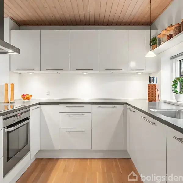 Moderne køkken med hvide skabe og stålgreb, en indbygget ovn og komfur. Træplanke-gulv og loft. Bordplade i mørkt materiale, med en vask og olie/eddike-sæt ved siden af. Vindue med planter.