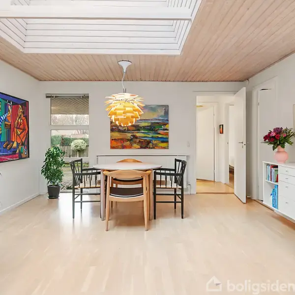 Spisebord med fire stole står i et lyst rum. Moderne lamper hænger fra loftet. Kunstværker pryder væggene, og planter dekorerer hjørnerne ved vinduerne.