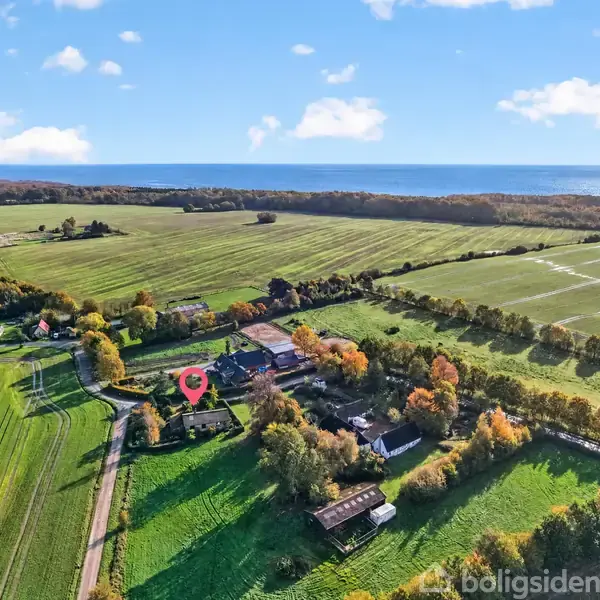Luftfoto viser en landsby omgivet af marker og skov, med hav i baggrunden. En rød markør peger på et hus blandt træer, inden for en landlig, frodig atmosfære.