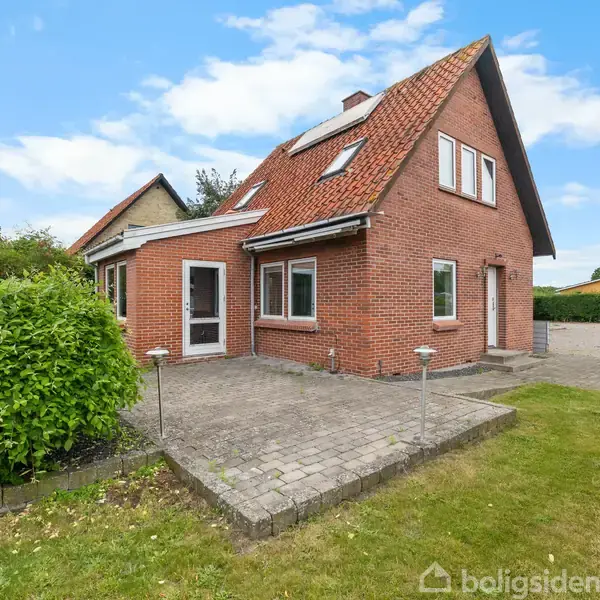 Et rødt murstenshus med skråt tag og flere vinduer står på en solskinsterrasse. Omkring huset er der en velholdt græsplæne og grønne buske under en blå himmel.