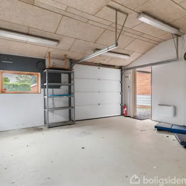 En garage med tomt gulv, to metalreoler og en hejseport. Lyse rør i loftet oplyser rummet. En rød brandslukker står ved døren, som åbner ud mod en murstenbygning.