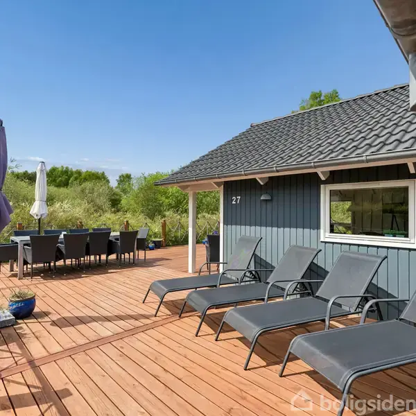 Blå træhus (objekt) med nummer 27, har solstole og spisebord på træterrasse (handling). Omkranset af grønt løv og blå himmel (kontekst).