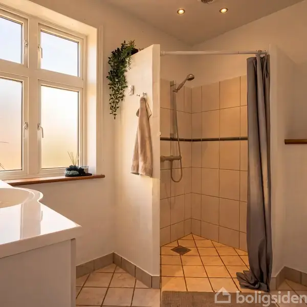 Badeværelse med bruser i hjørnet, beige fliser og bruseforhæng. Håndvask til venstre med spejl og planter. Toilet til højre. Sollys kommer ind gennem vinduet.