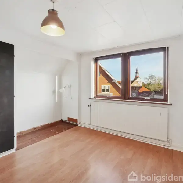 Et tomt værelse med trægulv, sort klædeskab og en enkelt hængelampe. Et stort vindue giver udsigt til et rødt tag og et kirketårn udenfor.