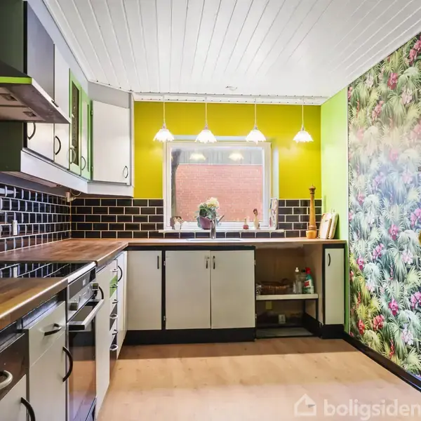 Køkken med hvide skabe og sort flisevæg, der forløber langs væggene. Tre hængelamper over en vask placeret foran et vindue. Højrevæggen har farverigt blomstertapet.