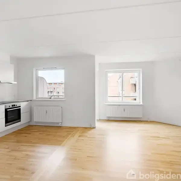 Et moderne køkken med hvide skabe og indbygget ovn i en lys, åben stue med trægulv. To vinduer giver udsigt til en anden bygning.