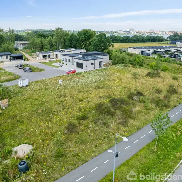 En tom grund ligger ubenyttet med vildtvoksende græs, omgivet af moderne huse i et forstadsområde. En asfalteret sti løber langs grunden, og biler er parkeret nær husene.