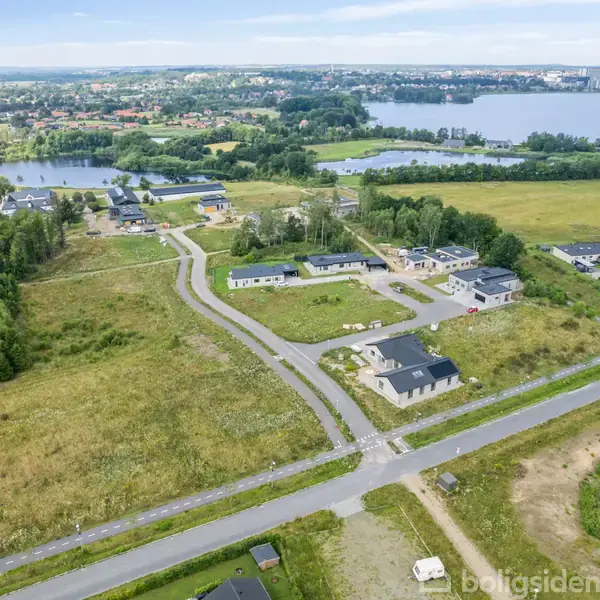 Landområde under udvikling med flere nye huse, omkranset af marker og skove. Veje forbinder bygningerne, og en sø samt en bydel ses i baggrunden.