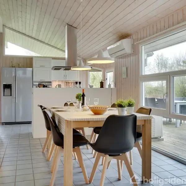 Spisebord med sorte stole står i en moderne spisestue. I baggrunden ses et lyst køkken. Stuen har store vinduer, der åbner ud mod en terrasse, omgivet af træer.