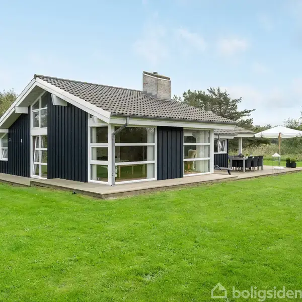 Et sort træsommerhus med store vinduer står i en frodig, grøn have. Der er en terrasse med havemøbler og en parasol ved siden af huset.