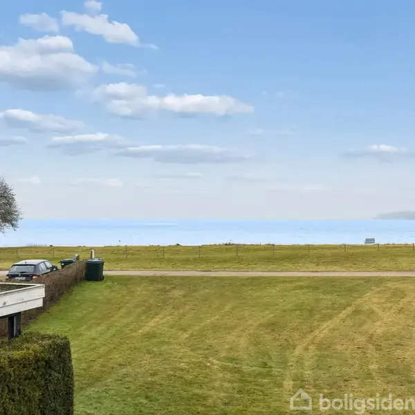 Græsplæne strækker sig mod en vej med en bil parkeret, omgivet af en hæk til venstre. I baggrunden ses havet under en blå himmel med lette skyer.