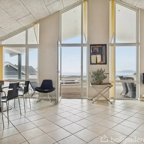 Spisestue med glasdøre åbne mod terrasse, moderne spisebord med sorte stole. Hængelampe lyser rummet, mens flisegulv og loftsdetaljer tilføjer elegance. Udsigt over havet gennem store vinduer.