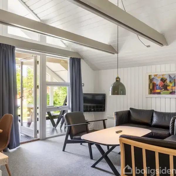 Stue med sort lædersofa omkring sofabord, lamper over bordet. Skydedøre åbner til altan med stole. Vægkunst til højre, tv i baggrunden. Lyse trævægge, synlige loftsbjælker.