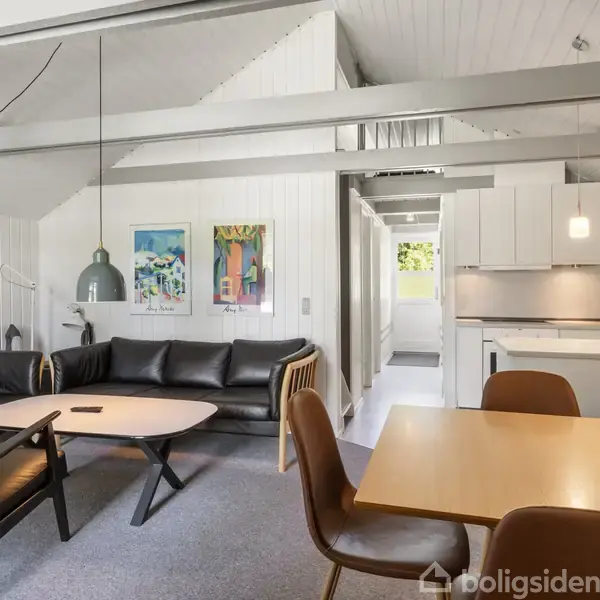 Stue med sort lædersofa og to stole omkring et lille sofabord. Et spisebord med fire stole står foran et moderne køkken. Lyse vægge og loft med malerier.