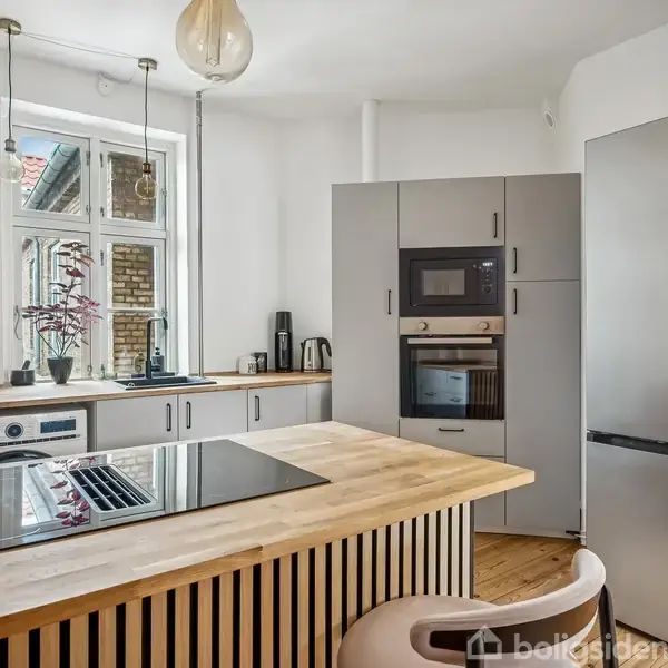 Køkken med gråt skabe og integreret ovn, mikrobølgeovn og køleskab. Kogeplade på træbordplade, højt vindue til venstre. Neutral indretning med moderne apparater og minimal dekoration.