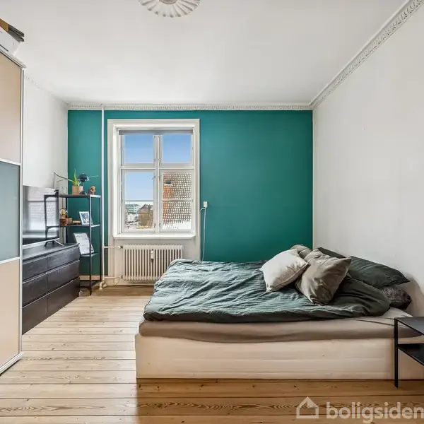 Et soveværelse med en dobbeltseng i midten, en skænk og hylder til venstre, altsammen på et trægulv. En grønmalet væg omkranser et vindue med udsigt.