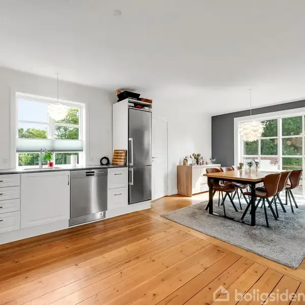 Køkken-alrum med hvidt køkken, stål hårde hvidevarer og trægulv. Spisebord med seks stole står på et tæppe ved glasdør, som giver udsigt til grøn have.