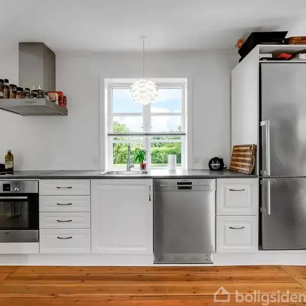 Moderne køkken med hvide skabe, rustfri stålapparater og træbordplade. Komfuret og opvaskemaskinen er indbygget, mens et stort vindue giver udsigt til grønne omgivelser. En hængende lampe lyser op.
