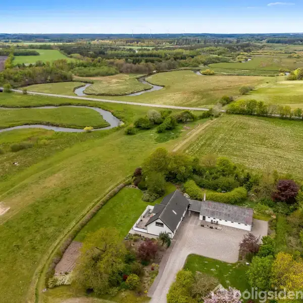 Et hvidt hus med gråt tag ligger ved siden af en stor grusbelagt indkørsel omgivet af grønt landskab med marker og snoede vandløb i baggrunden.