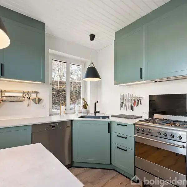 Køkken med grønne skabe og moderne stålkomfur. Vandhane ved hjørnevindue, ophængte køkkenredskaber og mørk loftlampe. Lys indretning med træ- og metaldetaljer.