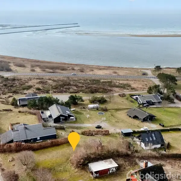 Hus set ovenfra med gult markeringsikon, omgivet af spredte huse og marker. I baggrunden ses en strandlinje og havet.