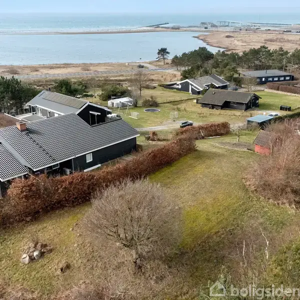Huse ligger spredt i et kystnært område med udsigt over havet. Et hus står centralt omgivet af buske og træer. Baggrunden viser en åben udsigt over vandet og skov.