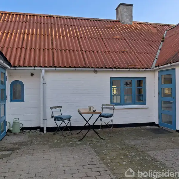 En gårdhave med hvide vægge og rødt tegltag. To metalstole står ved et lille bord med krus, omgivet af blå vinduer og døre på en brostensbelagt terrasse.