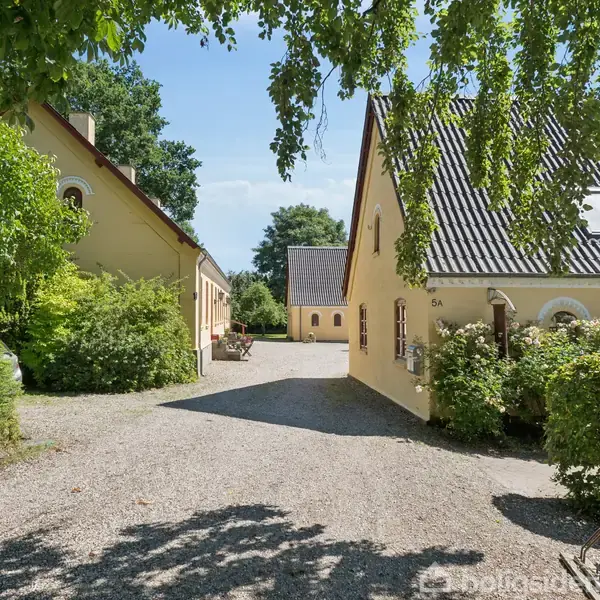 To gule huse står stille overfor hinanden i en solrig gård med grusvej. Store grønne træer og buske omgiver bygningerne. Text: "5A".
