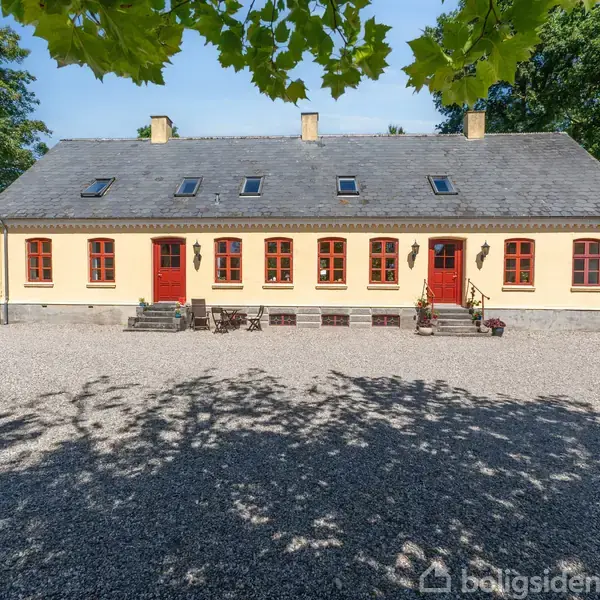 Et herregårdslignende hus med gul facade og røde døre står på en grusbelagt gårdsplads omgivet af træer. Skygger fra træerne falder over gruset.