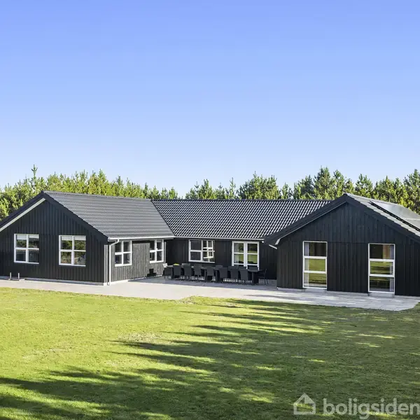 Sort træhus står stille i en åben, græsbeklædt have, omkranset af en række tætte træer under en klar blå himmel.