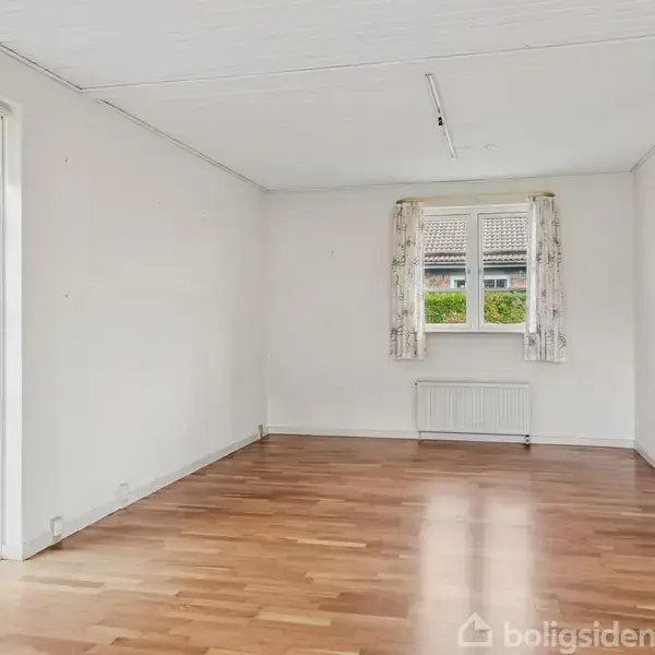 Et tomt rum med trægulv og hvide vægge. Rummet har to vinduer; det ene med gardiner og udsigt til en have, det andet med glasdør mod en terrasse.