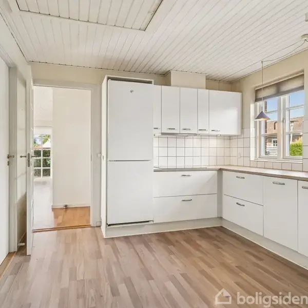 Et moderne køkken med hvidt inventar og trælaminatgulv. Køleskab, komfur og ovn indbygget. Store vinduer giver naturligt lys. Tilstødende rum ses gennem en åben døråbning.