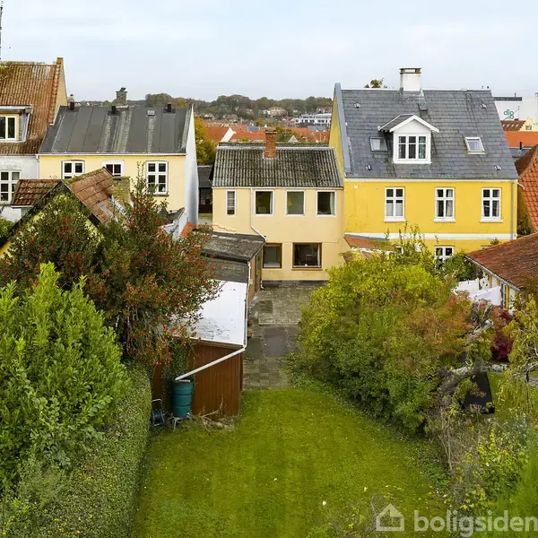 Flere huse står stille i række med velplejede haver foran sig. Et træskur og buske omkranser græsplænen, der strækker sig hen mod husene.
