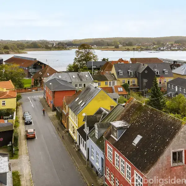 Farverige huse står langs en stille vej; en enkelt bil kører forbi. I baggrunden ligger en sø med frodige, grønne træer og kuperet terræn.