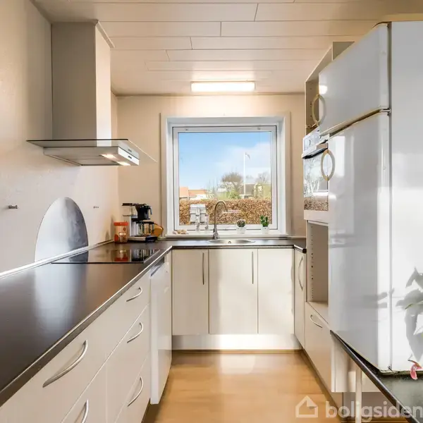 Et hvidt, moderne køkken med induktionskogeplade og emhætte til venstre, køleskab til højre, samt en kaffemaskine i vindueskarmen, har udsigt til en lys, grøn have.