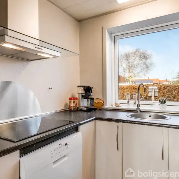 En moderne køkkenvask ved et stort vindue, omgivet af hvide skabe og en opvaskemaskine. En kogeplade og emhætte er også til stede, mens kaffemaskine og krydderier står ved siden af.